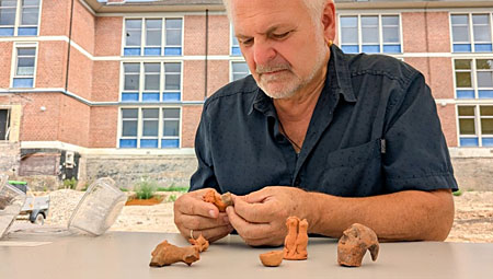 a man sits at a table in front of a modern brick-and-glass school building. He's handling a series of small clay figures, each about the size of his fingers.