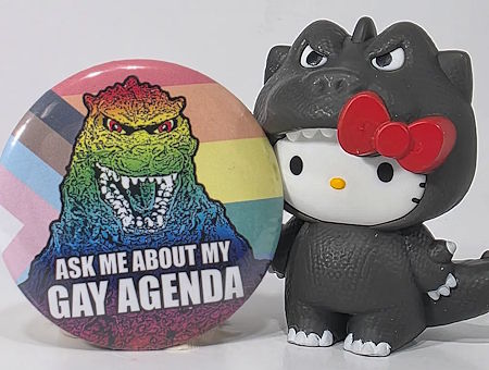 Hello Kitty Godzilla holding up a pin featuring rainbow Godzilla in front of the Pride flag, reading ASK ME ABOUT MY GAY AGENDA.