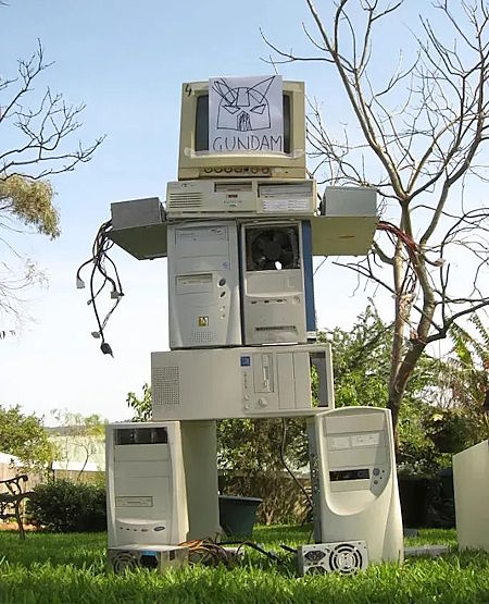 [A humanoid robot structure constructed from stacked, obsolete desktop computer towers and a CRT monitor, standing outdoors on a grassy lawn. Loose wires hang from the computer cases acting as arms. Taped to the monitor ''head'' is a piece of paper with a hand-drawn picture of a mecha helmet (from the Mobile Suit Gundam franchise).] ''GUNDAM''