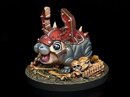 image of a painted miniature of a smiling Moo Deng as a Khorne Berzerker. She wears spiked red and bronze armor with metal horns and stands on a base littered with skulls and bones.