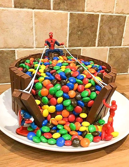 A homemade cake with Kit-Kat bars standing vertically around the outside, and M&Ms spilling out of a gap in the wall. A Spider-Man figure sits on top, with two webs shooting out to grab the bars that are falling over. Iron Man helps lift one of them, and Captain America helps lift the other.